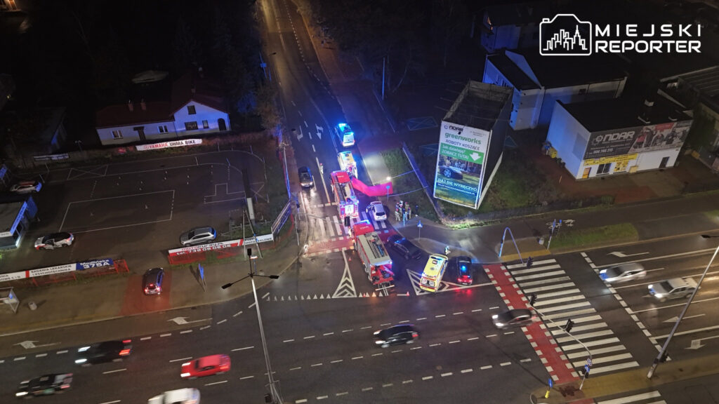 Służby ratunkowe i radiowozy stoją na skrzyżowaniu przy przejściu dla pieszych, otoczone niebieskimi światłami.