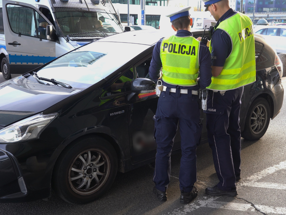 Dwóch policjantów w odblaskowych kamizelkach kontroluje kierowcę czarnego samochodu obok radiowozu.