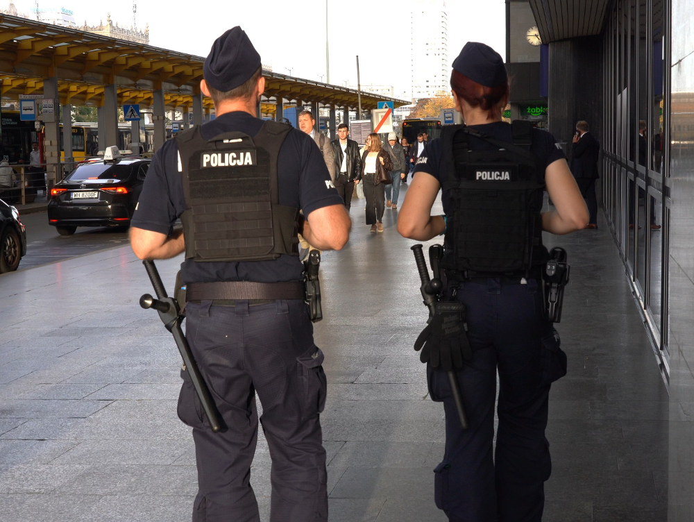 Dwóch umundurowanych policjantów z pałkami patroluje chodnik przy ulicy, wśród przechodniów i samochodów.