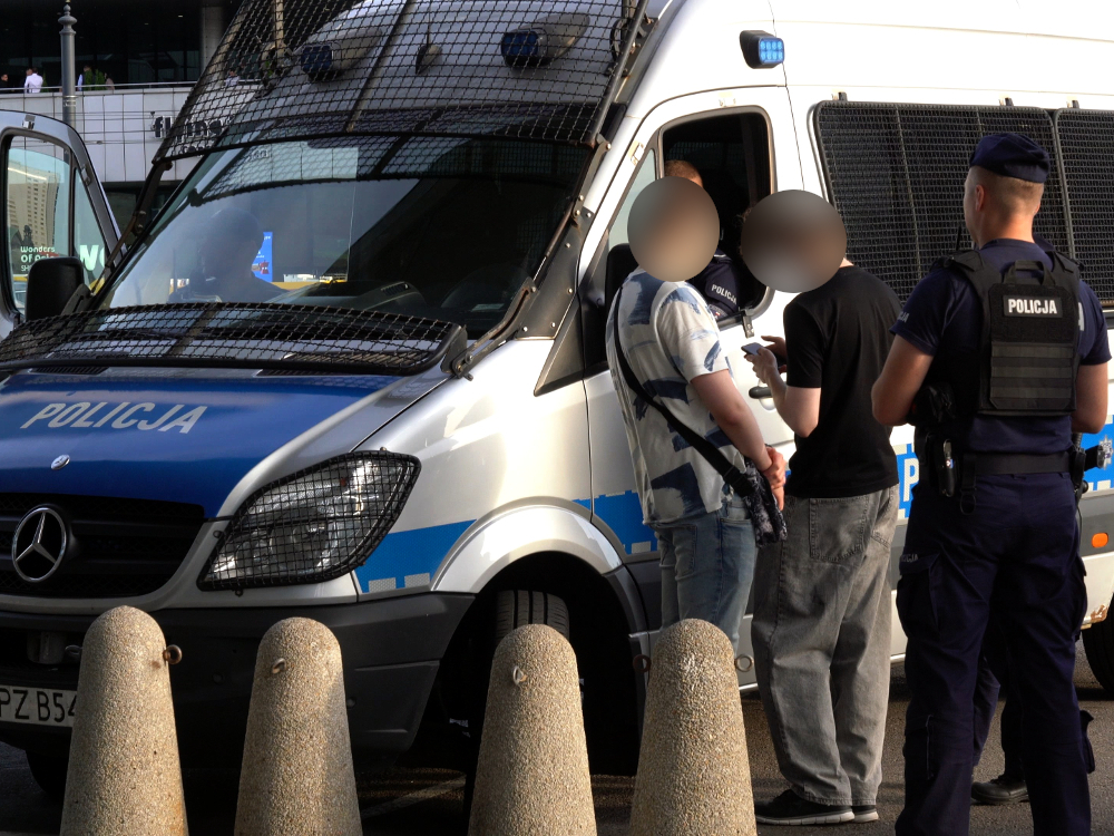 Policjant stoi obok dwóch mężczyzn przy policyjnym busie na ulicy.