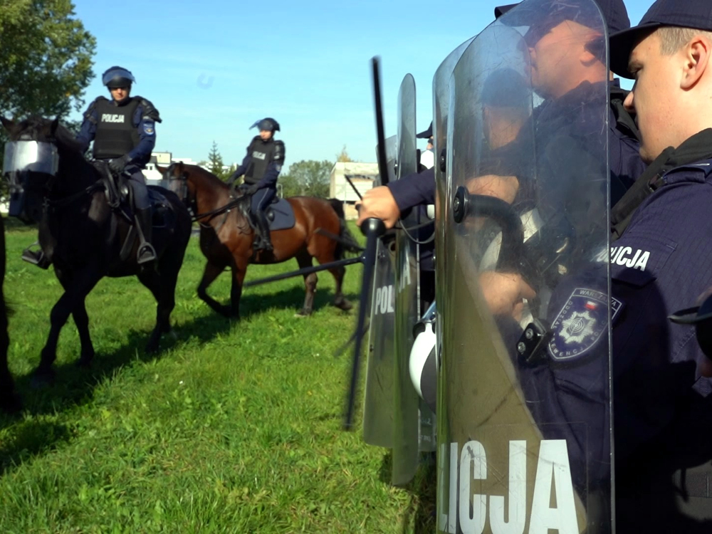 Policjanci na koniach i funkcjonariusze z tarczami ćwiczą na trawiastym terenie