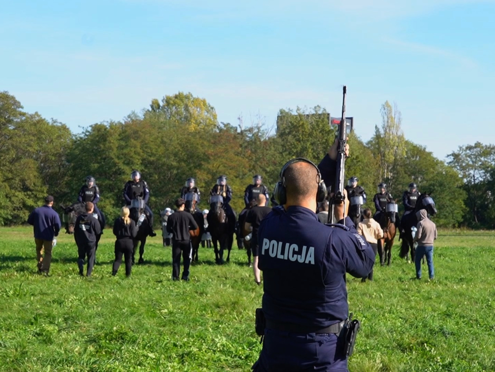 Policjant z długą bronią obserwuje funkcjonariuszy na koniach i grupę osób na otwartej łące
