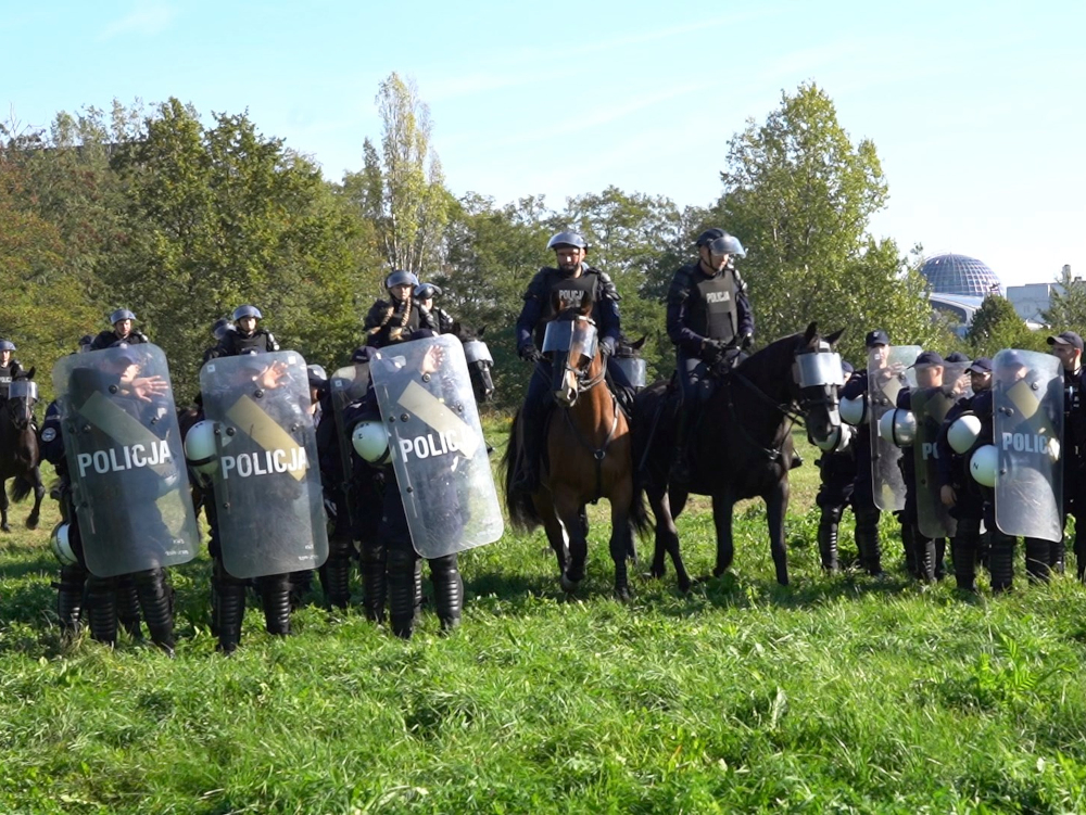 Policjanci w kaskach i z tarczami „POLICJA” stoją w szyku, a za nimi siedzą funkcjonariusze na koniach.