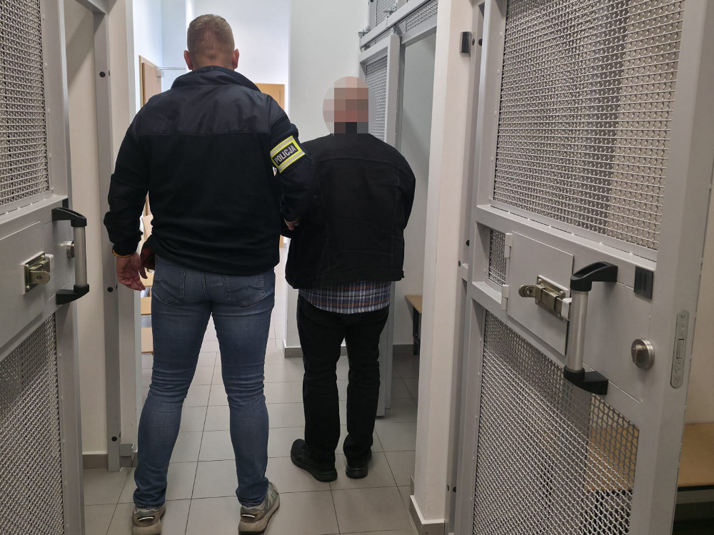 Funkcjonariusz policji prowadzi mężczyznę korytarzem między otwartymi, metalowymi drzwiami.