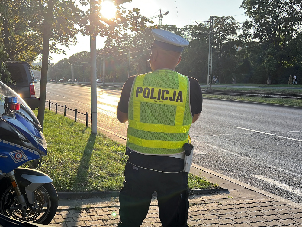 Policjant w kamizelce z napisem „POLICJA” stoi przy drodze, obok oznakowanego policyjnego motocykla.