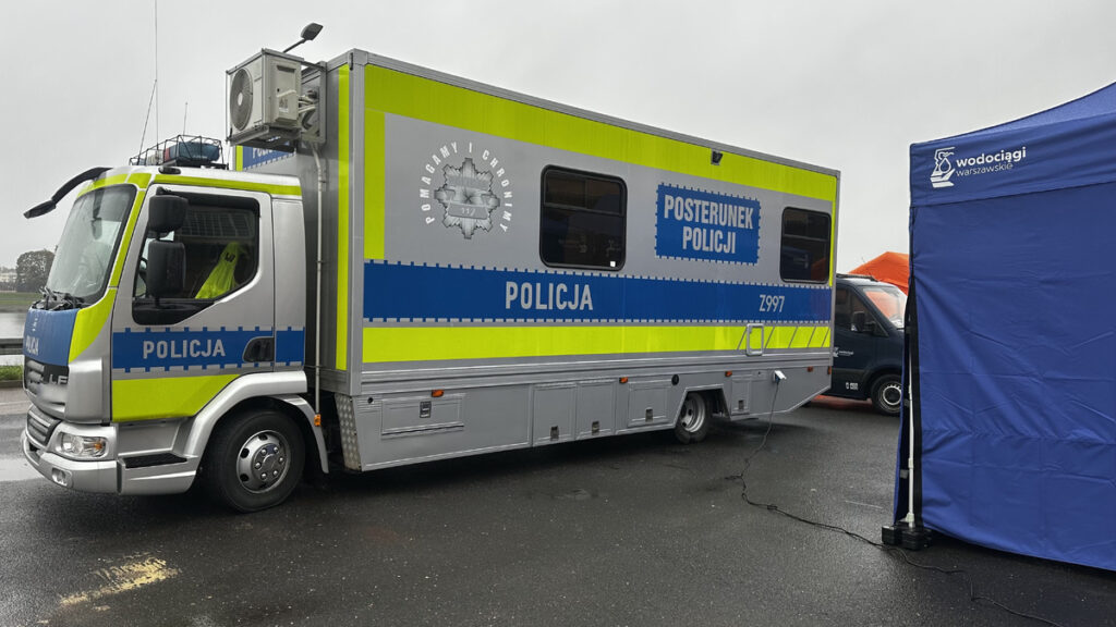 Policyjny mobilny posterunek stoi na mokrym parkingu obok niebieskiego namiotu z napisem „Wodociągi Warszawskie”.