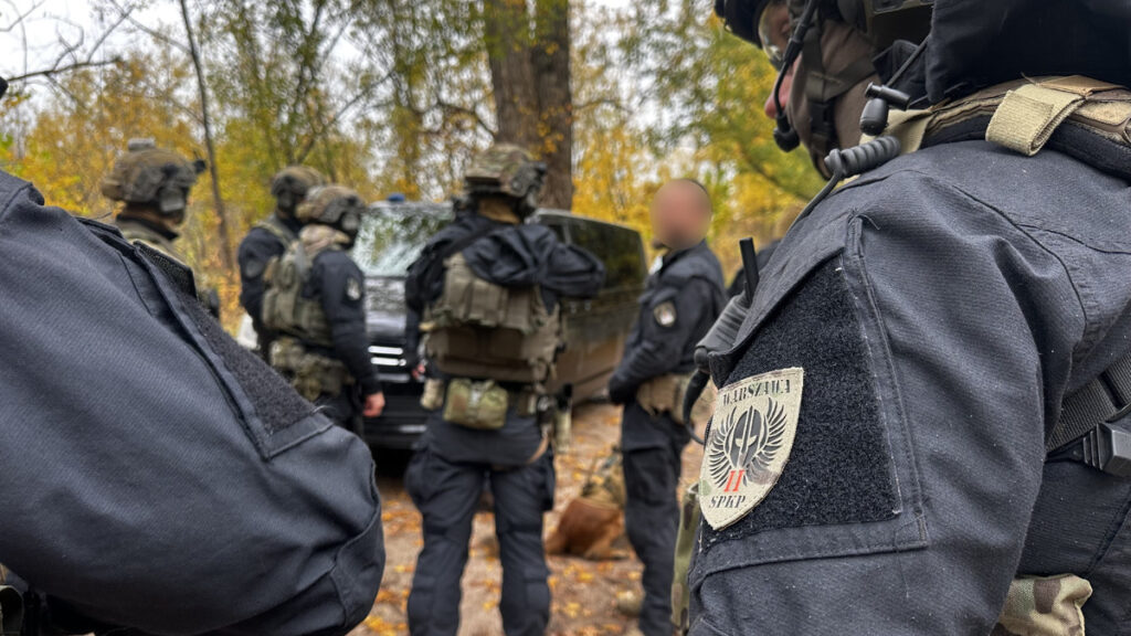 Uzbrojeni funkcjonariusze w mundurach i hełmach stoją przy wojskowym pojeździe wśród jesiennych drzew.
