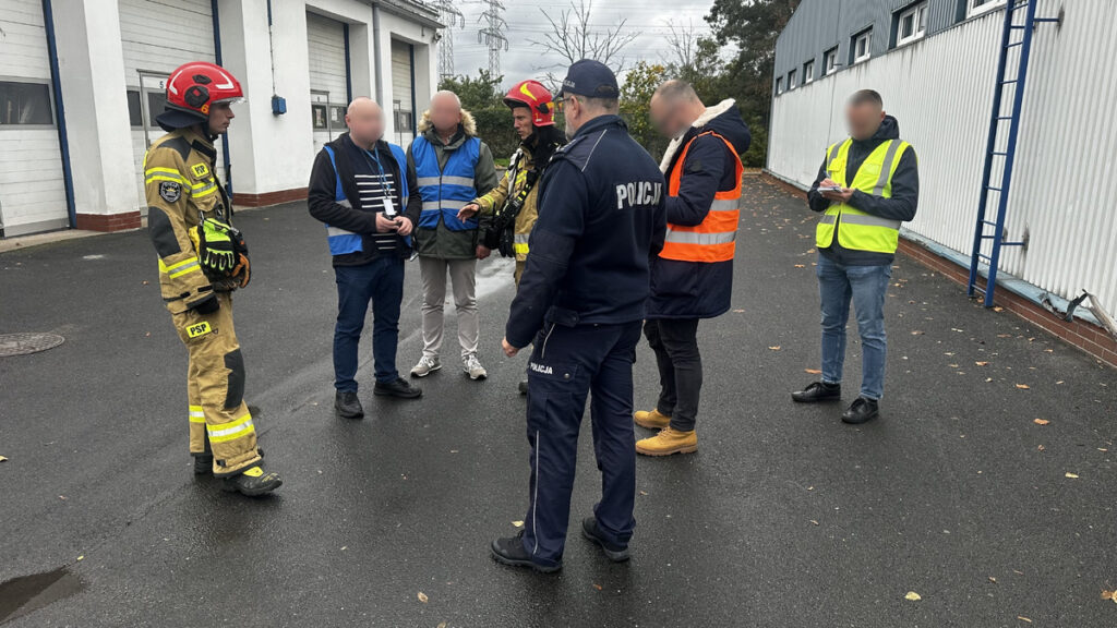 Policjant, strażacy i osoby w kamizelkach odblaskowych rozmawiają na terenie między budynkami przemysłowymi.