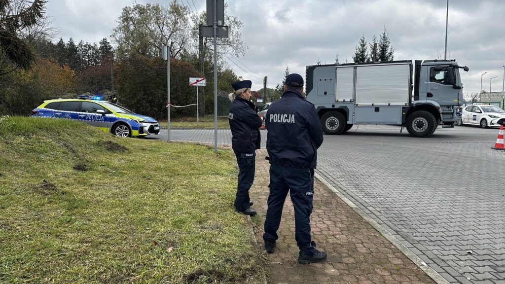 Dwóch policjantów stoi przy drodze, obok radiowozu z włączonymi światłami i ciężarówki służb ratunkowych.