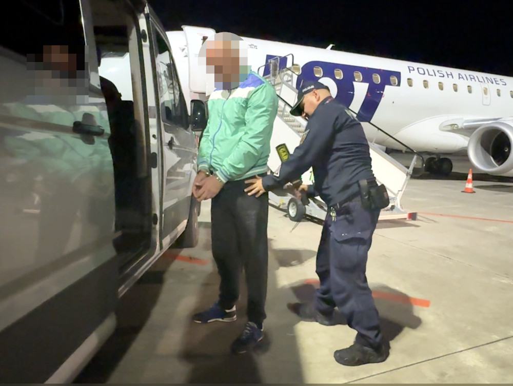 Funkcjonariusz eskortuje skutego mężczyznę do samochodu na płycie lotniska, obok samolotu Polskich Linii Lotniczych LOT.