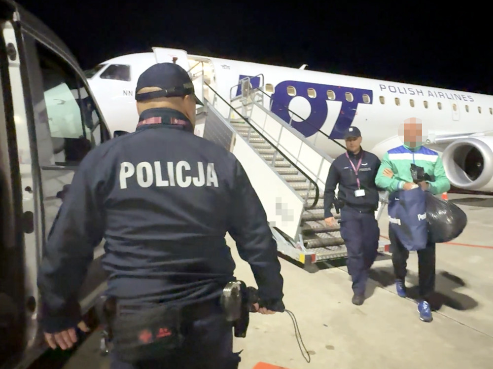 Policjant eskortuje zakutego w kajdanki mężczyznę w stronę samochodu przy schodach samolotu pasażerskiego LOT.