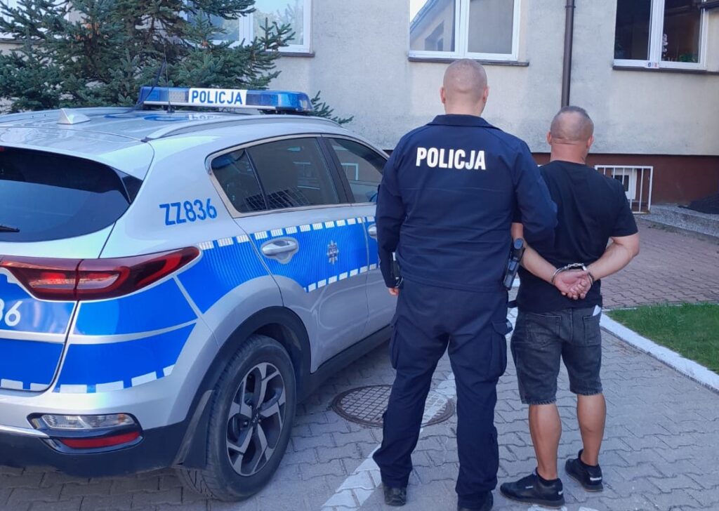 Policjant prowadzi skutego mężczyznę obok oznakowanego radiowozu na parkingu przy budynku.