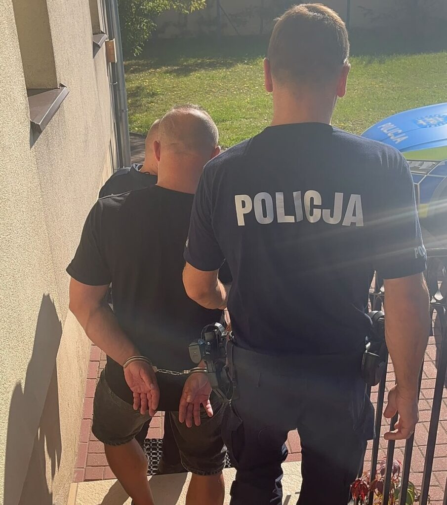 Policjant prowadzi skutego kajdankami mężczyznę wzdłuż budynku, obok radiowozu.