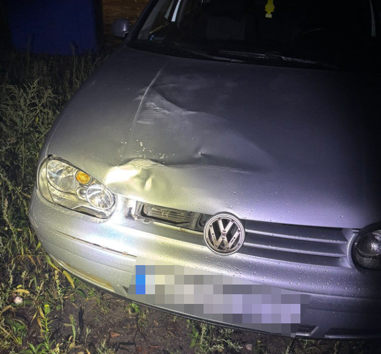 Srebrny Volkswagen z wgniecioną maską i uszkodzonym przednim zderzakiem stoi przy zarośniętym poboczu nocą.