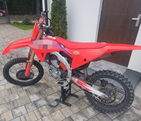 Czerwony motocykl terenowy stoi na podstawce na wyłożonym kostką podjeździe przy wejściu do budynku.