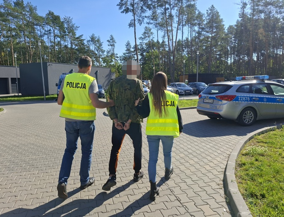 Policjanci prowadzą zatrzymanego mężczyznę w kajdankach w stronę radiowozu na parkingu.