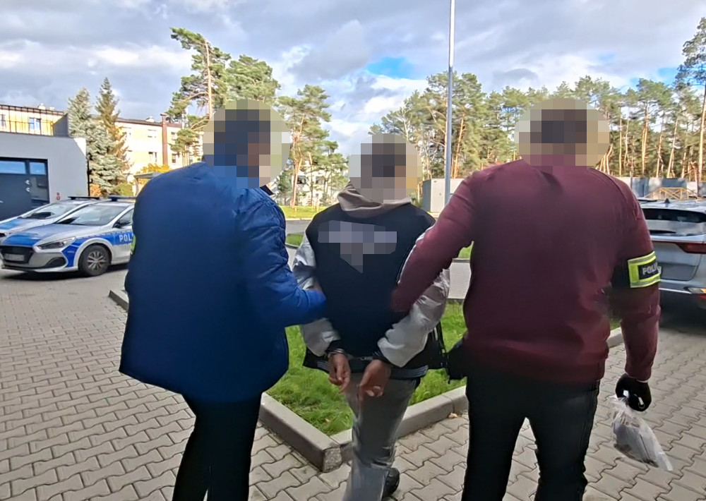 Dwóch funkcjonariuszy prowadzi mężczyznę w kajdankach po parkingu, obok zaparkowanego radiowozu policyjnego.