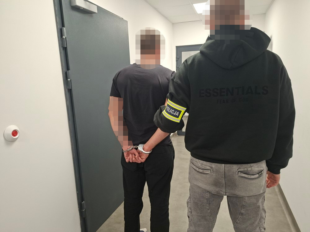 Policjant prowadzi skutego kajdankami mężczyznę korytarzem budynku.