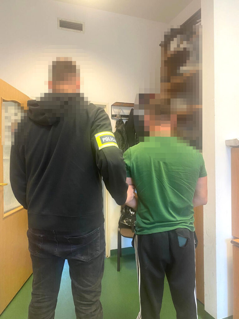 Policjant prowadzi zatrzymanego mężczyznę korytarzem budynku; twarze obu osób są zamazane.
