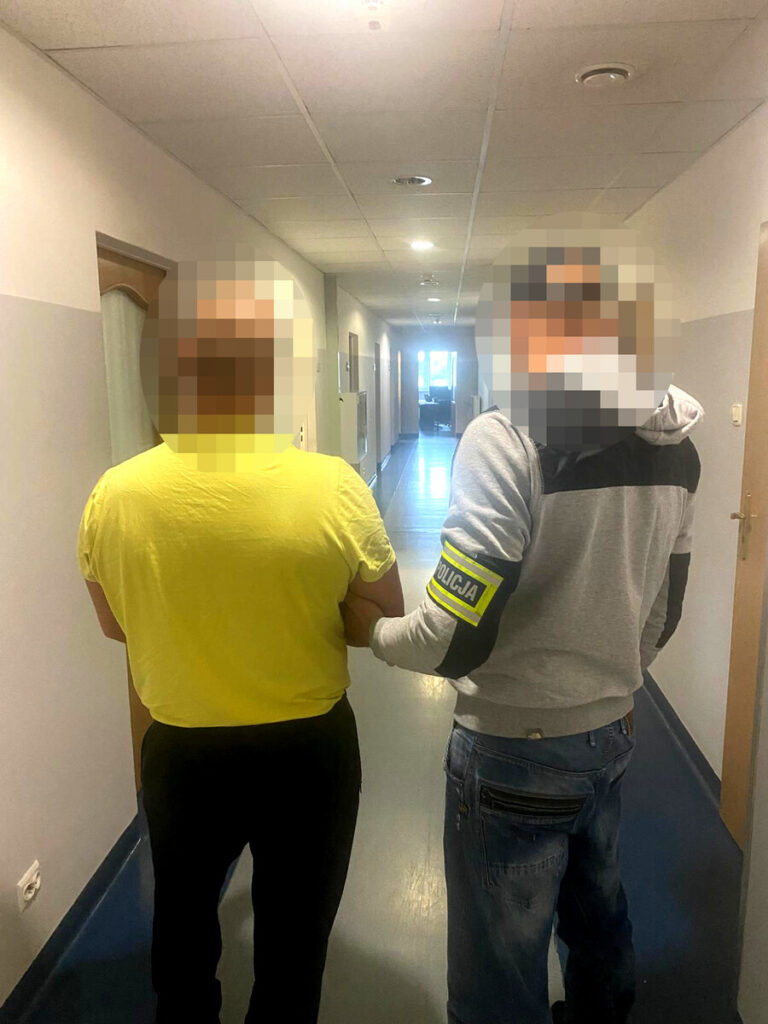 Policjant prowadzi zamaskowanego mężczyznę korytarzem budynku; twarz mężczyzny jest zamazana.