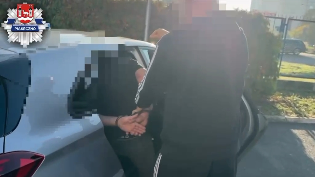 Policjant prowadzi zakutego w kajdanki mężczyznę obok zaparkowanego samochodu.