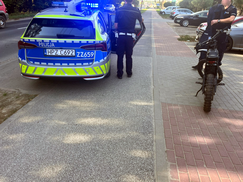 Policjant stoi przy radiowozie z włączonymi niebieskimi światłami, obok funkcjonariusz przy motocyklu na chodniku.