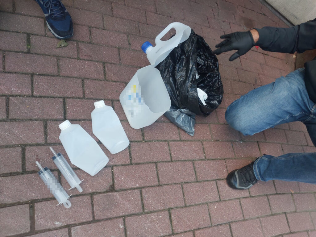 Plastikowe kanistry, strzykawki i czarny worek leżą na chodniku, obok nich klęczy osoba w rękawiczkach.