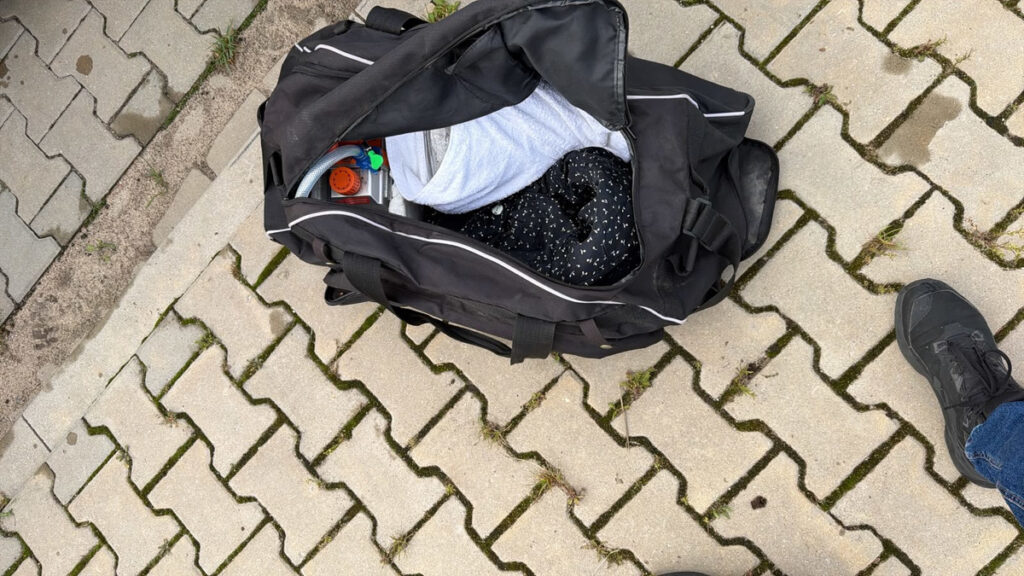 Otwarta czarna torba z ubraniami i przedmiotami leży na brukowanym chodniku obok buta.