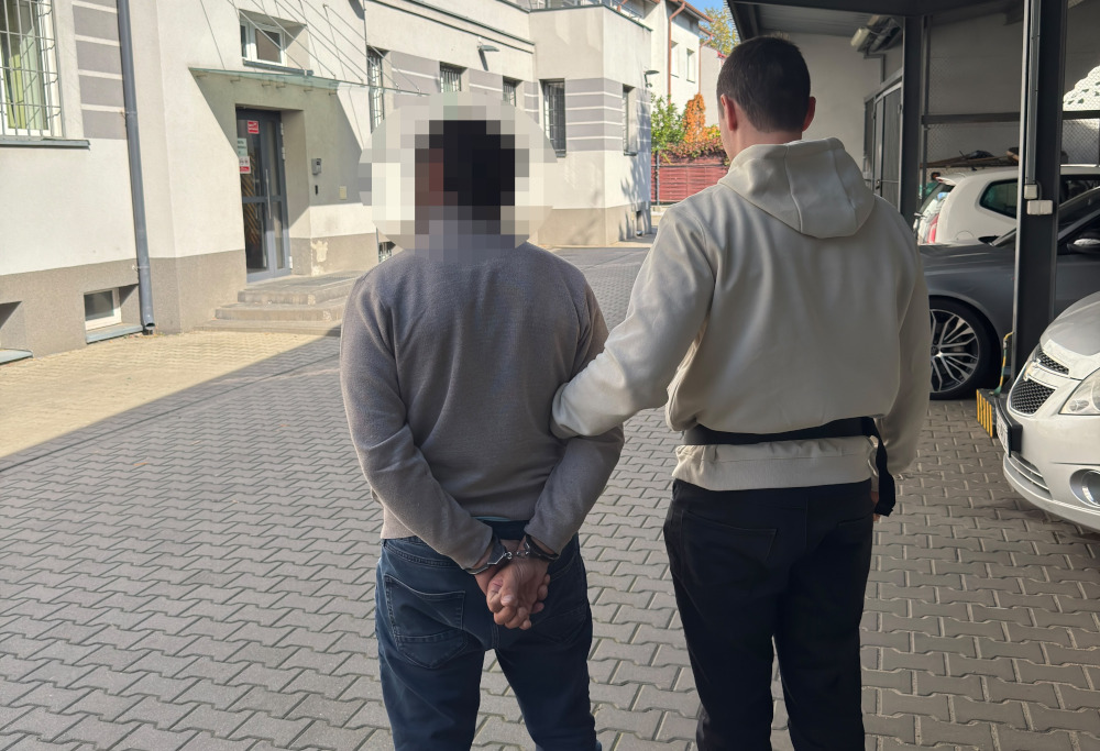 Policjant prowadzi mężczyznę z rękami skutymi kajdankami za plecami przed budynkiem i zaparkowanymi samochodami.