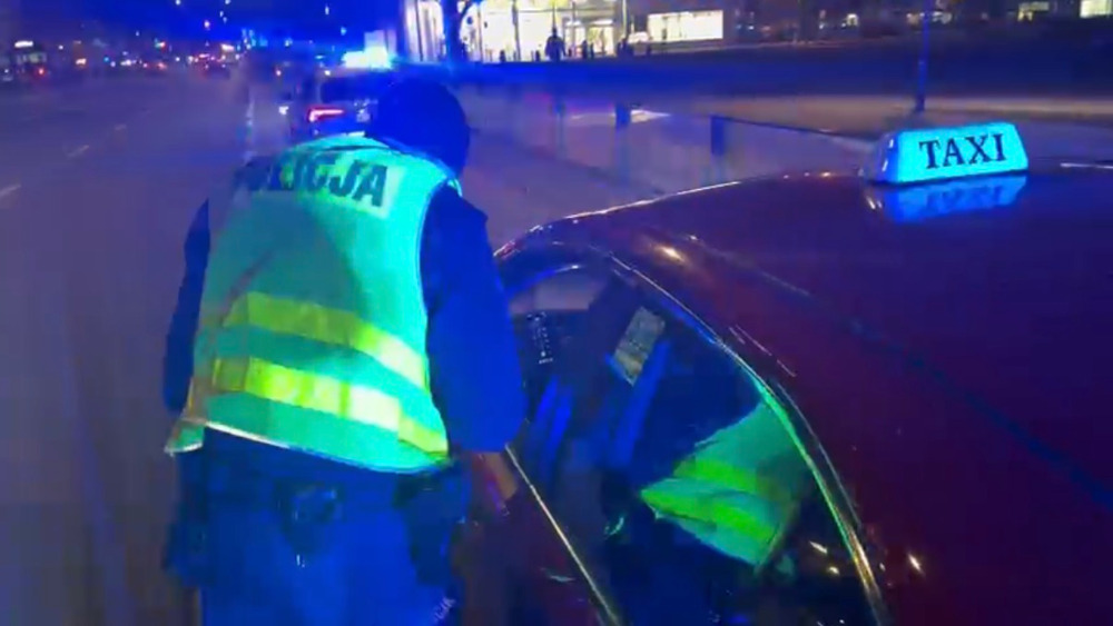 Policjant w odblaskowej kamizelce zagląda do taksówki zatrzymanej nocą przy oświetlonej ulicy.