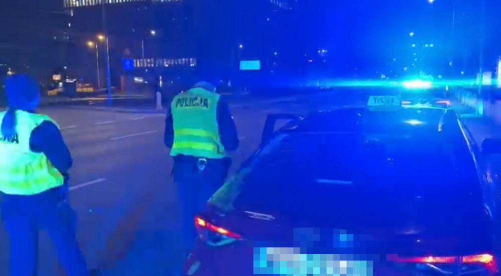 Policjanci w odblaskowych kamizelkach kontrolują samochód na nocnej ulicy przy włączonych niebieskich światłach radiowozu.