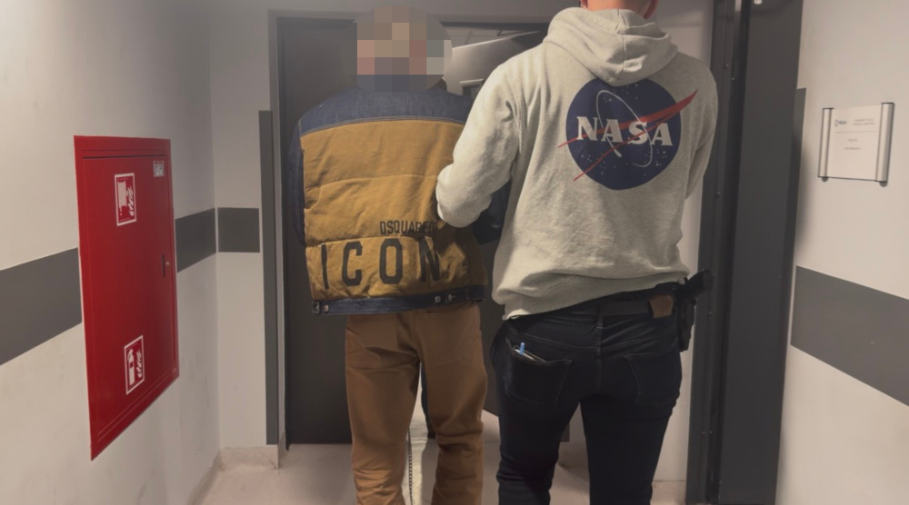 Policjant w kamizelce prowadzi mężczyznę z zasłoniętą twarzą przez korytarz budynku.