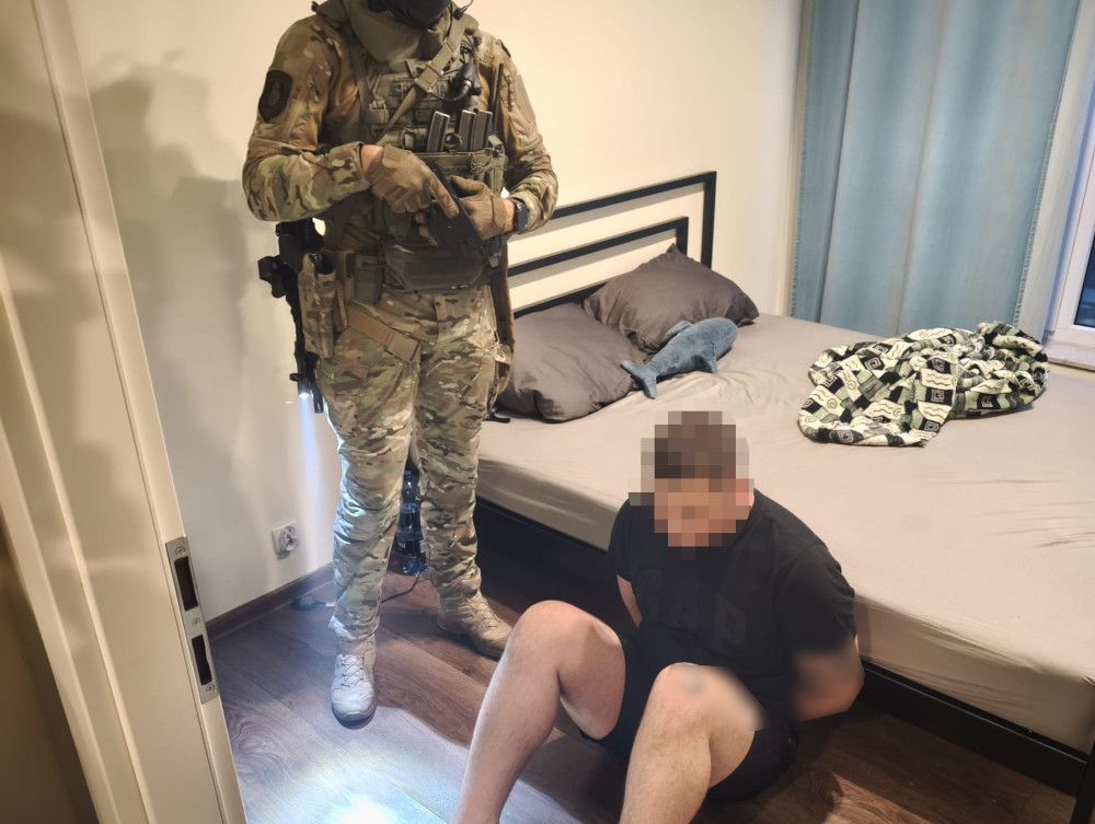 Uzbrojony funkcjonariusz stoi obok siedzącego na podłodze mężczyzny w sypialni przy łóżku.