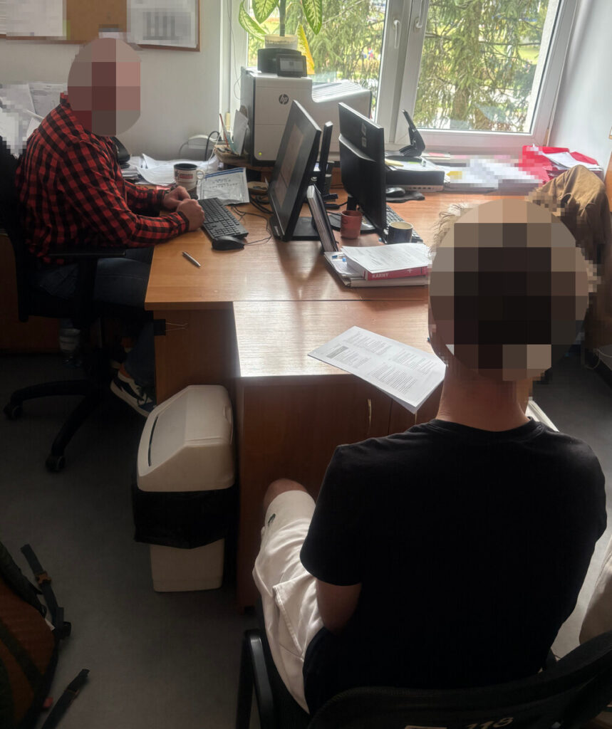 Mężczyzna w czarnej koszulce siedzi naprzeciw pracownika przy biurku z komputerami w biurze.