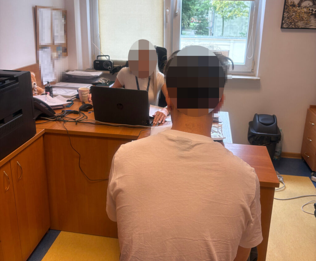 Mężczyzna siedzi tyłem do aparatu przy biurku, naprzeciwko kobiety pracującej przy laptopie w pokoju biurowym.