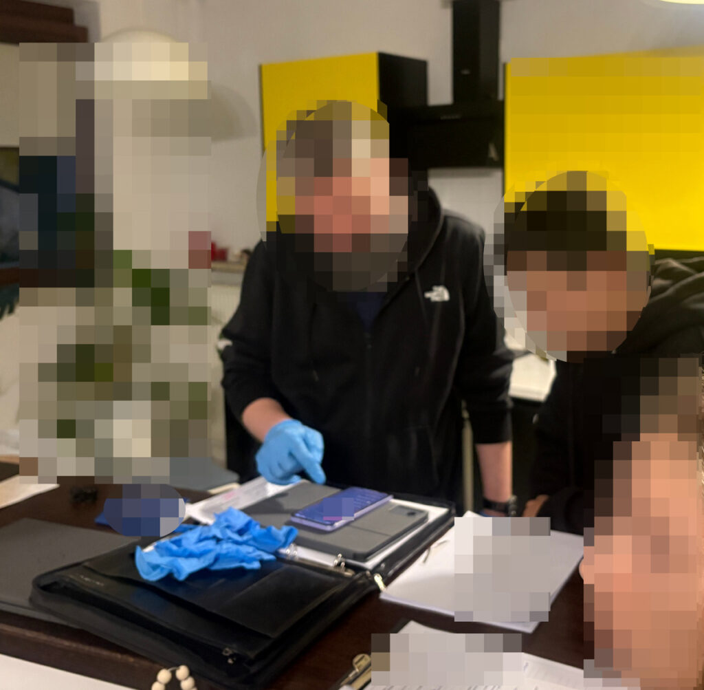 Mężczyźni w rękawiczkach oglądają telefon i dokumenty na stole w pomieszczeniu kuchennym.