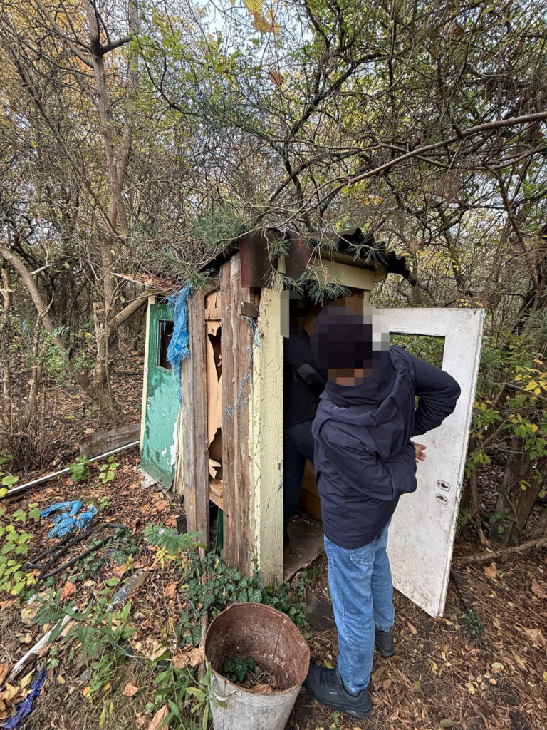 Mężczyzna w ciemnej kurtce stoi przy wejściu do zniszczonej drewnianej budki w zarośniętym lesie.