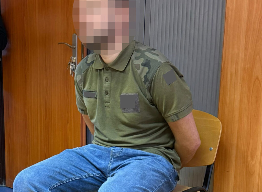Mężczyzna w zielonej koszulce moro siedzi na krześle w pomieszczeniu, z zamazaną twarzą i rękami za plecami.