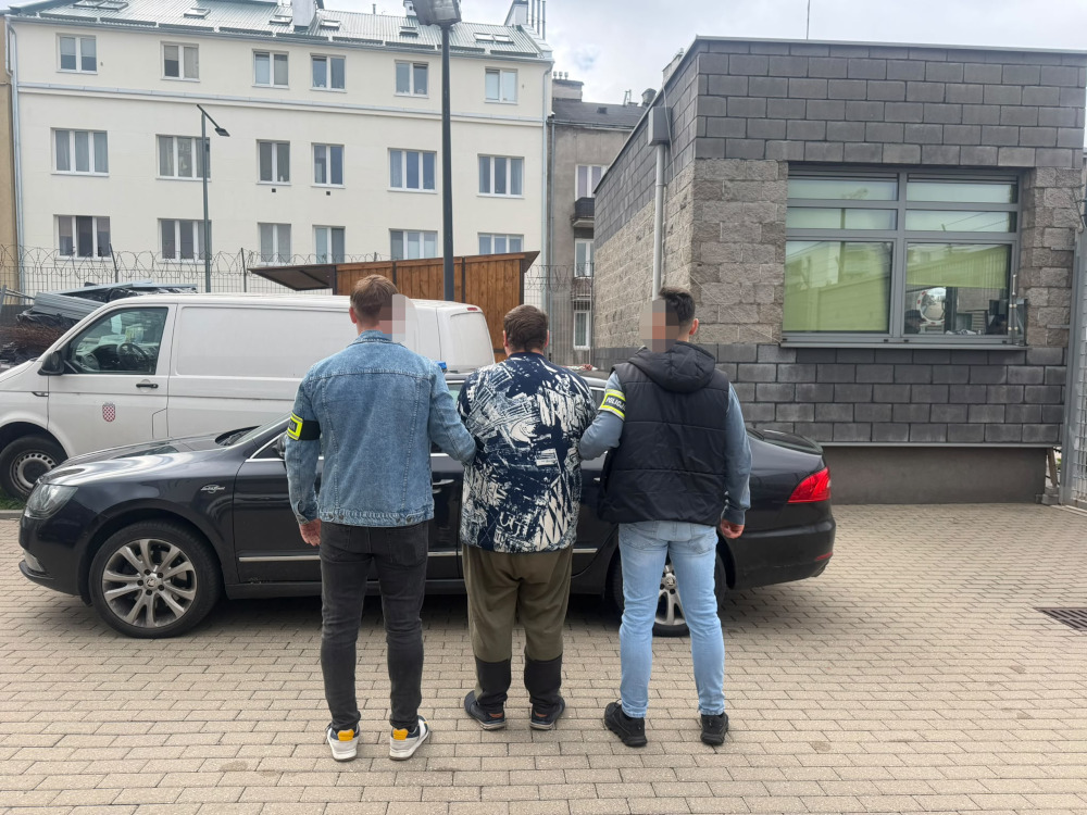 Trzech mężczyzn, dwóch z oznaczeniami na rękawach, stoi tyłem przy czarnym samochodzie na parkingu.