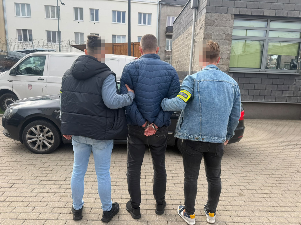 Mężczyzna z rękami skutymi z tyłu stoi między dwoma osobami w kamizelkach z oznaczeniami służb, obok zaparkowanych aut.