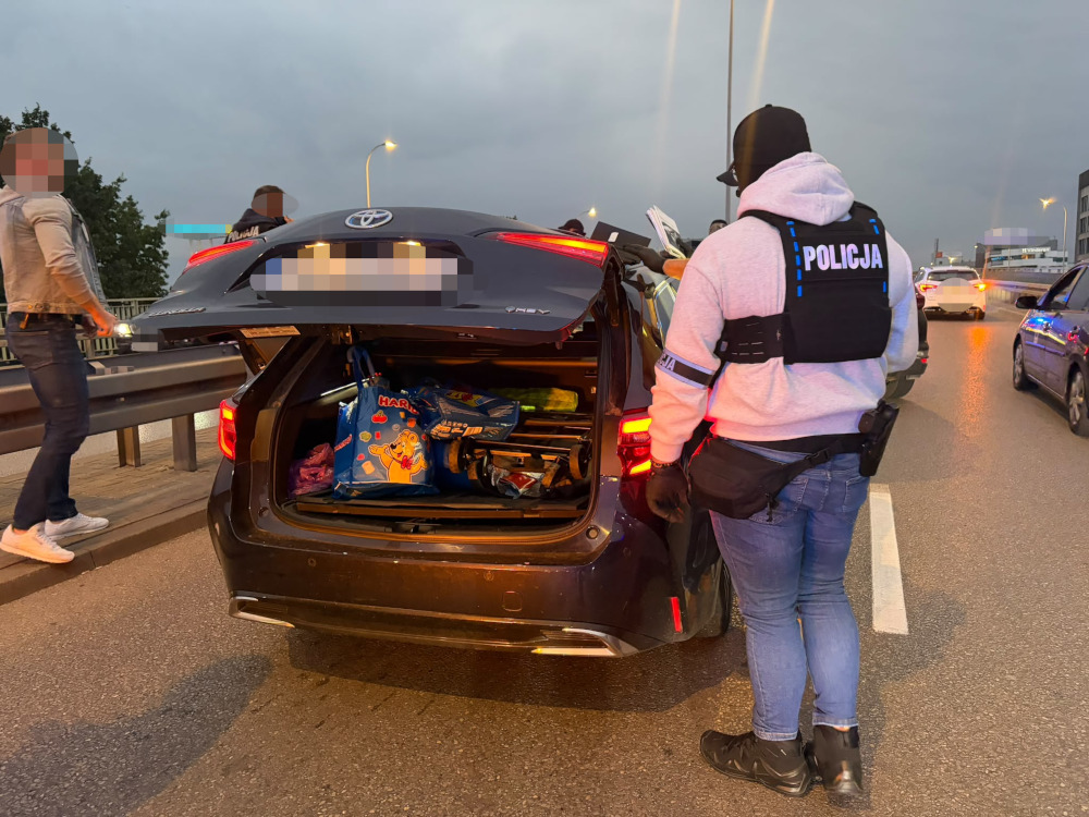 Policjant w kamizelce z napisem „POLICJA” stoi przy otwartym bagażniku Toyoty na drodze.