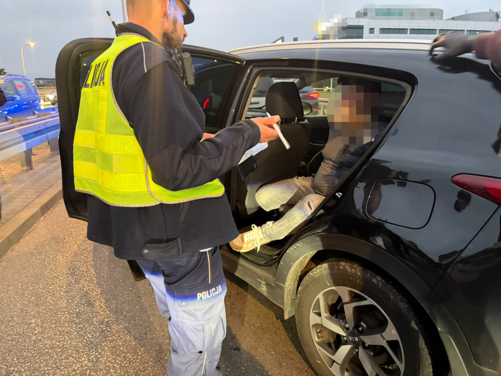 Policjant w żółtej kamizelce sprawdza dokumenty mężczyzny siedzącego w otwartym samochodzie na poboczu drogi.