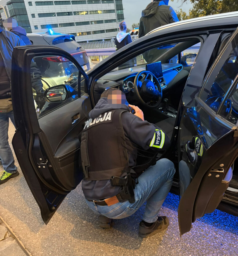 Policjant w kamizelce z napisem „POLICJA” sprawdza wnętrze otwartego samochodu, obok stoi radiowóz z włączonymi sygnałami