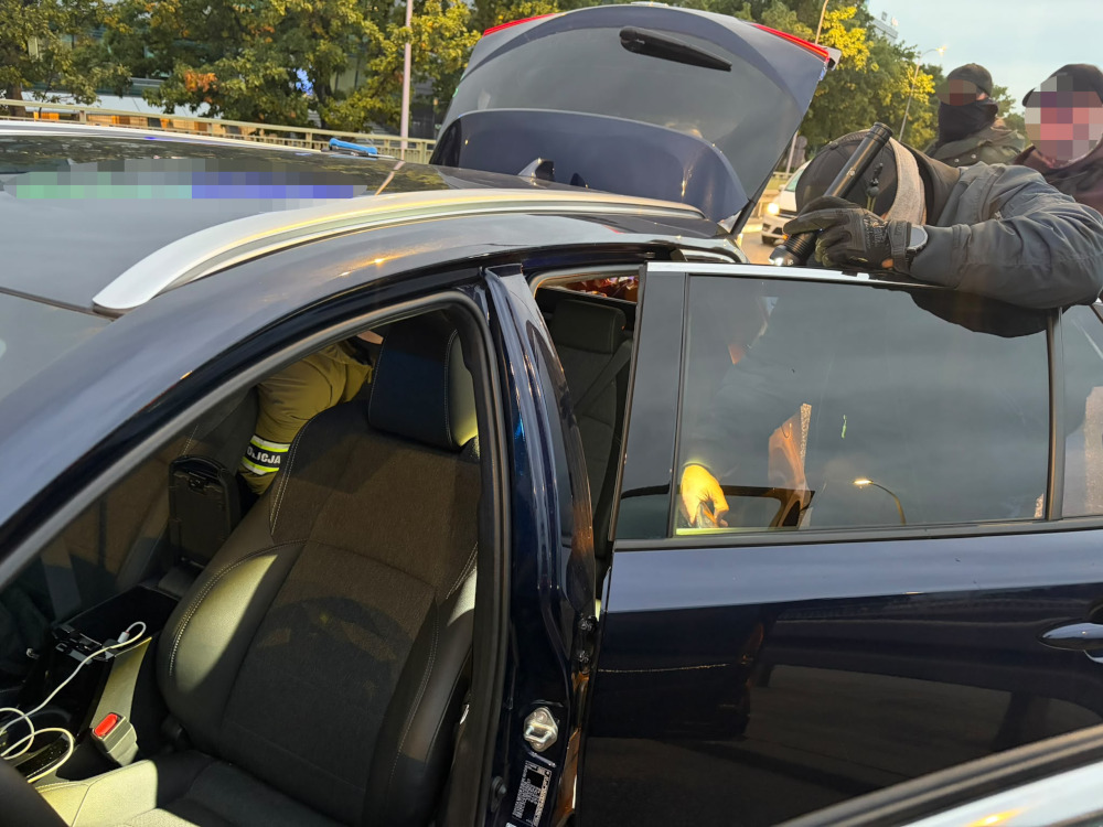 Umundurowani funkcjonariusze przeszukują granatowy samochód z otwartymi drzwiami i bagażnikiem na parkingu.