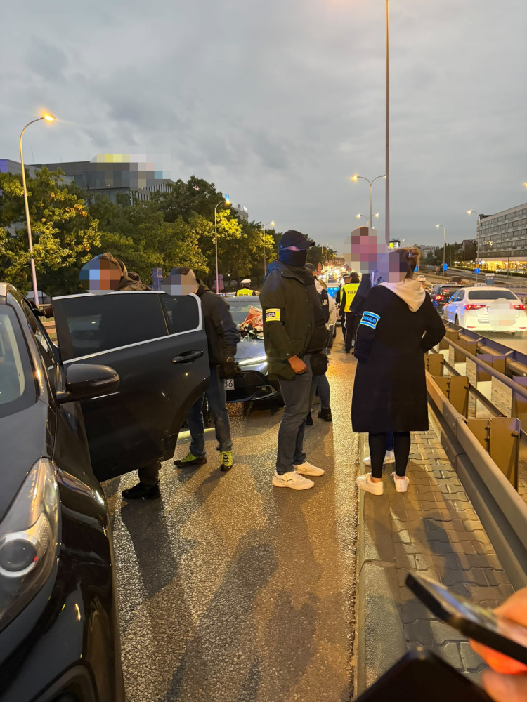 Policjanci i funkcjonariusze w cywilnych ubraniach stoją przy otwartym samochodzie na miejskiej ulicy o zmierzchu