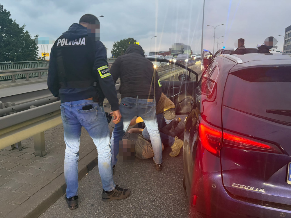 Policjanci zatrzymują mężczyznę obok Toyoty na poboczu drogi, przy barierze ochronnej i ruchu ulicznym.