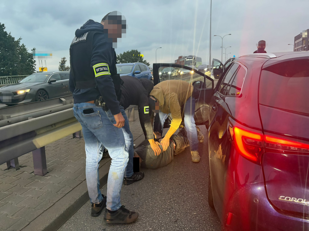 Funkcjonariusze w cywilnych ubraniach obezwładniają mężczyznę na jezdni obok czerwonej Toyoty Corolla.