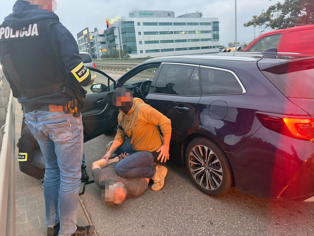 Policjant i mężczyzna w pomarańczowej bluzie obezwładniają osobę przy otwartych drzwiach ciemnego samochodu na parkingu.