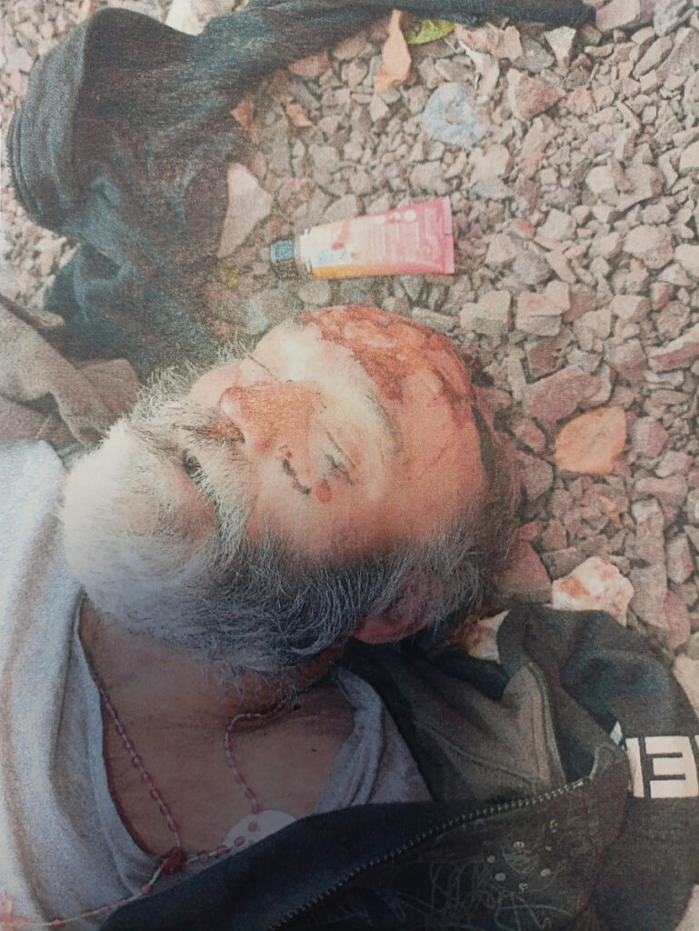 Siwowłosy mężczyzna leży na żwirze, z widocznymi obrażeniami twarzy; obok leżą fragmenty ciemnego ubrania i opakowanie.