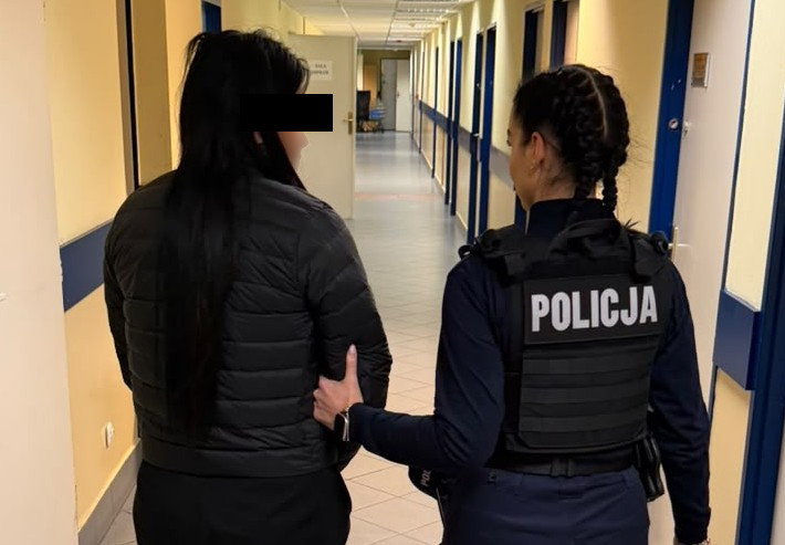 Policjantka prowadzi kobietę korytarzem w budynku; na kamizelce funkcjonariuszki widnieje napis „POLICJA”.